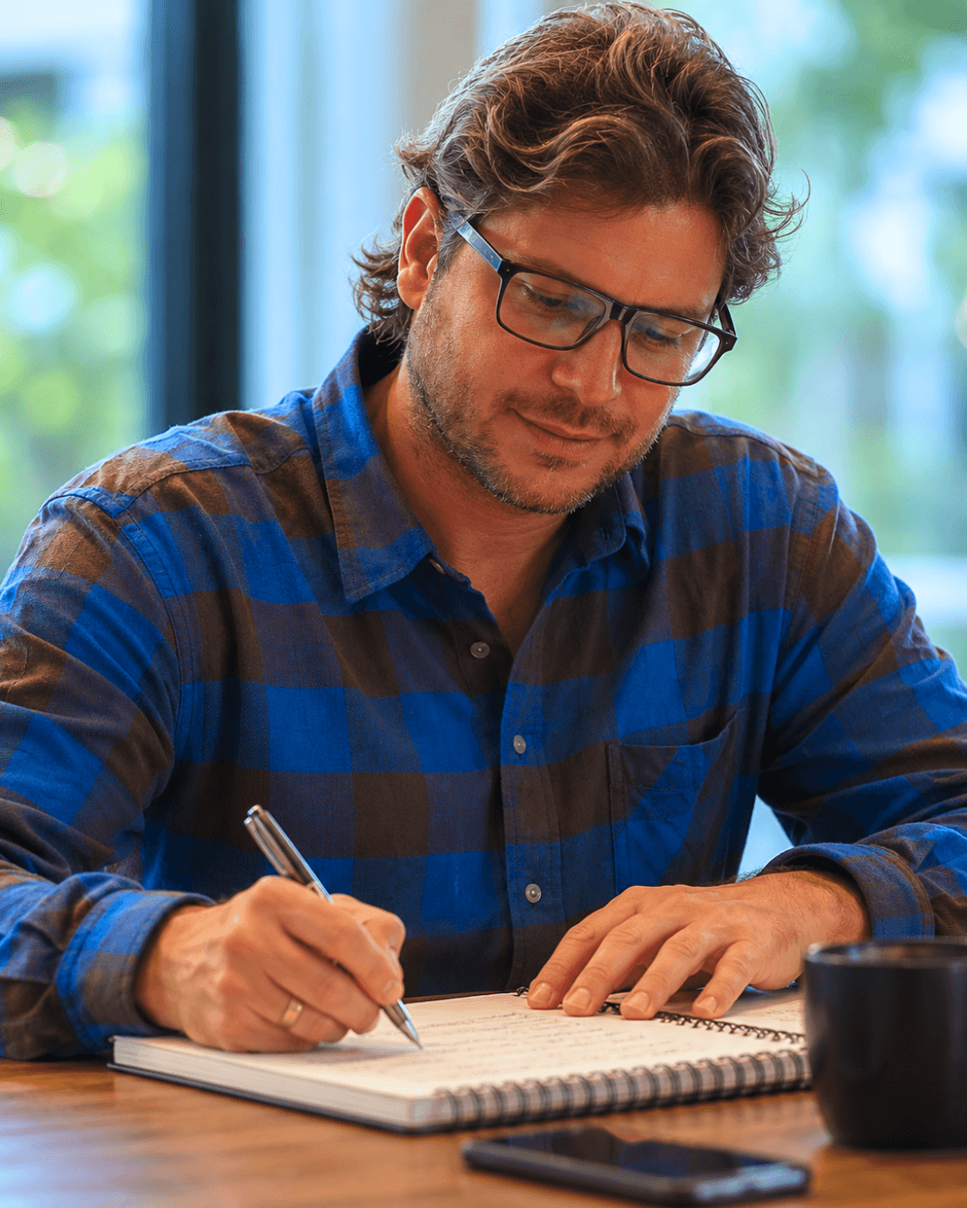 Lucas Carballo escribiendo en su cuaderno con anteojos y camisa a cuadros azules, en una mesa de madera con luz natural detrás.